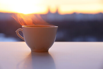 Hot coffee or tea cup with smoke on sunshine background. View from the window to silhouettes of city buildings