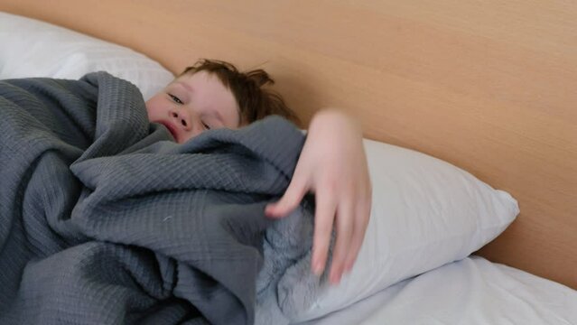 A sad boy is lying on the bed with a fever and a headache, and he has a headache. Covered with a blanket over his head. He looks out from under the blanket and discontentedly throws a soft toy into th