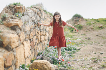 Cute caucasian child girl in a summer dress walks barefoot over stones and wild flowers. Summer holidays, childhood and natural wild plants and flowers among the stones