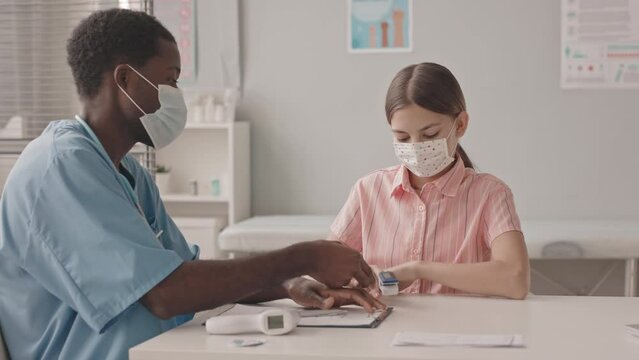 Medium slowmo shot of 13-year-old Caucasian girl being examined by young African-American doctor with pulse oximeter and contactless infrared thermometer