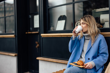 Hungry caucasian woman in blue jacket, drinking her takeaway coffee, before eating.