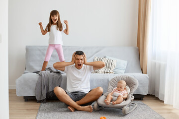 Indoor shot of sad upset depressed man wearing white casual style T-shirt and jeans short sitting on floor with his children, covering ears, being tired of screaming kids.