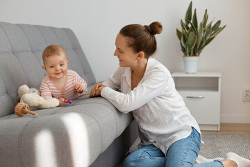 Positive young adult woman with bun hairstyle wearing white shirt and jeans sitting on floor and playing with crawling kid, mother playing with daughter, maternity leave.