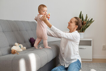 Portrait of delighted woman wearing white shirt and jeans sitting on floor and holding her infant baby wearing striped sleeper, mother playing with her little daughter.