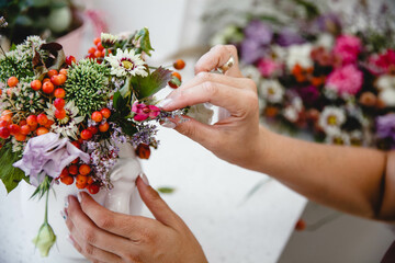 A florist makes a bouquet of different flowers and rose hips in a white vase at a white table in his workshop