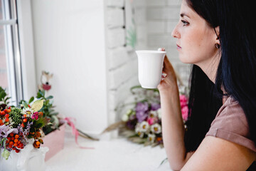 A young brunty European woman with long hair sits at a table with flowers and drinks coffee from a white mug