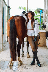 Vertical nice horsewoman cowgirl, equestrienne, hat, breeches lean on. Horse back riding. Horse animal care washing in paddock, tendering after ride. Outdoor wooden farm, ranch, yard. Countryside