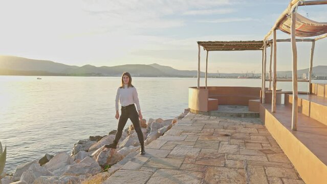 Carefree young woman plays by the sea while waiting during golden hour. UHD quality