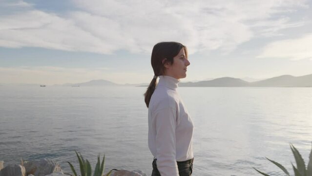 Medium truck shot of a woman walking on a pier during golden hour and gazing upon the sea. UHD quality