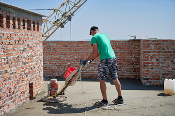 Full length of man builder using troweling machine while screeding floor in new building under blue sky. Male worker finishing concrete surface with floor screed grinder machine at construction site.