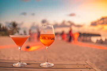 Two cocktail drinks with blur beach party people and colorful sunset sky in the background. Luxury outdoor leisure lifestyle, relaxing and romantic colors, blurred people partying on a summer evening - obrazy, fototapety, plakaty