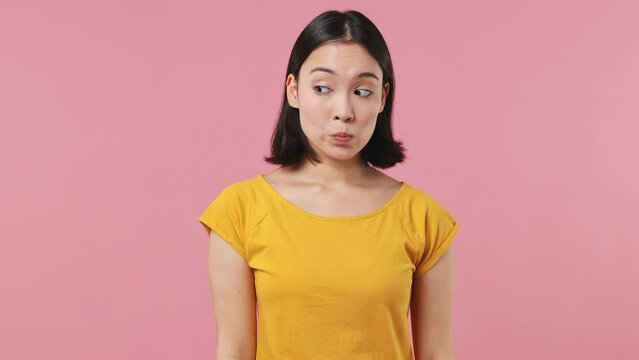 Confused shy shamed young woman of Asian ethnicity 20s wear yellow t-shirt look camera spreading hands say oops ouch oh omg i am so sorry isolated on plain pastel light pink background studio portrait