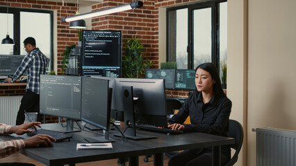 Programer with clipboard sitting down at desk in front of programer writing code while team of...
