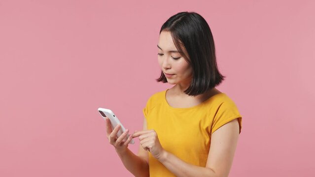 Young woman of Asian ethnicity 20s wears yellow t-shirt hold use mobile cell phone browsing swipe chatting send sms doing online shopping isolated on plain pastel light pink background studio portrait
