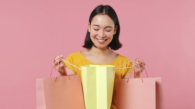 Charming young woman of Asian ethnicity 20s years old wear yellow t-shirt holding looking into package bags with purchases after shopping isolated on plain pastel light pink background studio portrait