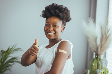 Young african american woman showing thumbs up and her arm with band aid after coronavirus Covid-19...