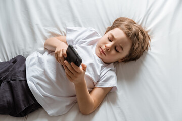Obraz na płótnie Canvas Little baby boy lays on his bed and plays with a smartphone. Close-up portrait. Holds in hands black cell phone. Concept of online education. White room and clothes. Telephone user. Save child vision