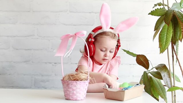 Cute little girl with bunny ears on Easter vacation. A child with headphones listens to cheerful music and puts Easter colored eggs in the basket. Baby dressed as a rabbit. Happy Easter