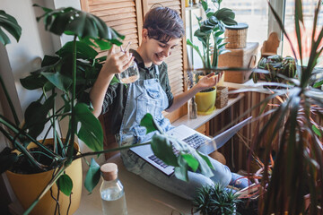 Happy young girl with laptop surrounded by home plants on the balcony, online communication, plant...