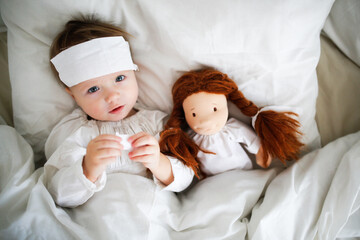 Cute baby with a doll with a compress on his head, a sick child in bed, bed rest during illness....