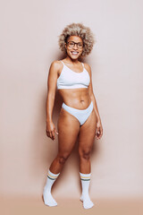 Cheerful beautiful afro latin american woman in white underwear and eyeglasses isolated on beige background at studio. Stretch marks and cellulite on the skin. Real people, real body, natural beauty. - obrazy, fototapety, plakaty