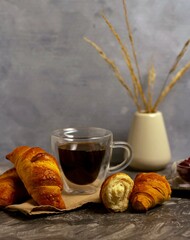 Croissants with coffee in a transparent glass on a gray background. Nearby is a vase with dried flowers.