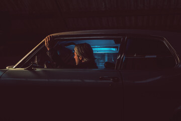 Man with blond hair sits inside an American classic muscle car with opened window at night.
