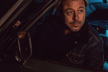 Man with blond hair sits inside an American classic muscle car with opened door and window at night.