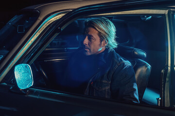 Man with blond hair sits inside an American classic muscle car with opened door and window at night.