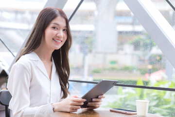 Young Asian business woman is looking at camera in her hands tablet or notepad while she is sitting at coffee shop among business background in the city.