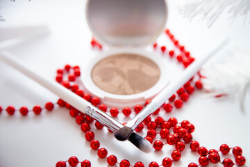 A white pillbox with powder and bronzer lies on a white background, thin makeup brushes and bright red beads lie in front. High quality photo