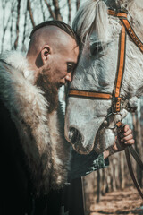 A medieval warrior Viking with a beard and a muscular physique poses in the forest with a horse