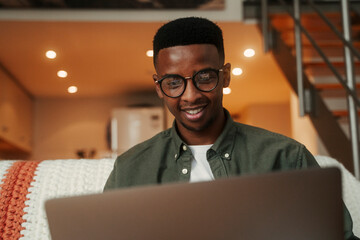 African American male sitting on sofa smiling while typing on laptop organising work for university 