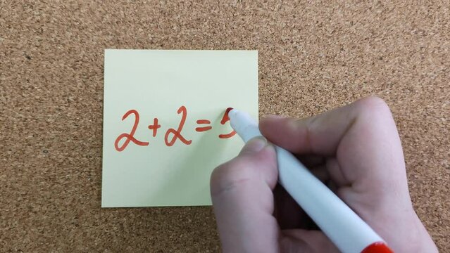 2+2=5. An arithmetic equation with an error. A simple mathematical equation on a yellow sticker. A woman's hand with a red marker writing. The concept of teamwork, synergy.