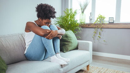 Young woman in gray clothes is sitting curled up on bed at home. Upset African American girl is...