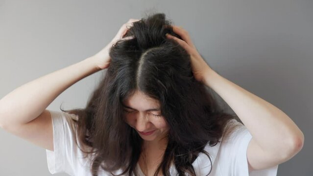 Portrait of a young Caucasian woman, strongly scratching her head with dark tangled hair. Gray background. The concept of dandruff and itchy scalp.
