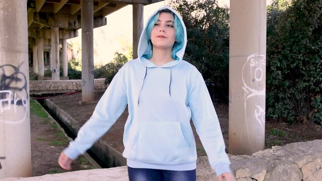 Teenager in light blue oversize hoodie and jeans Smiling and dancing. Blue haired teen girl stays outdoors against bridge pillars. Clothing mockup