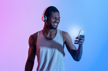 Handsome sporty black guy with smartphone wearing headset, choosing music playlist in neon light