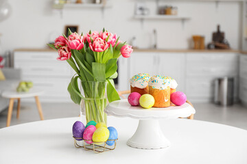 Stand with Easter cakes, eggs and tulips on dining table in kitchen