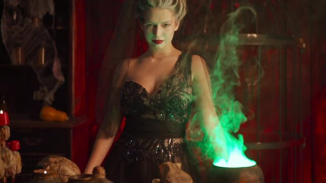 Halloween witch with cauldron. Beautiful young woman flowers in hair conjuring, making witchcraft. Standing gothic dark room. Enchantress prepare love potion use magic spell. Slow motion Handheld 