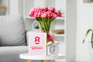 Beautiful bouquet of fresh flowers with alarm clock and greeting card on table in room. International Women's Day celebration