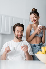 happy bearded man using smartphone near pleased woman in bra holding cup.