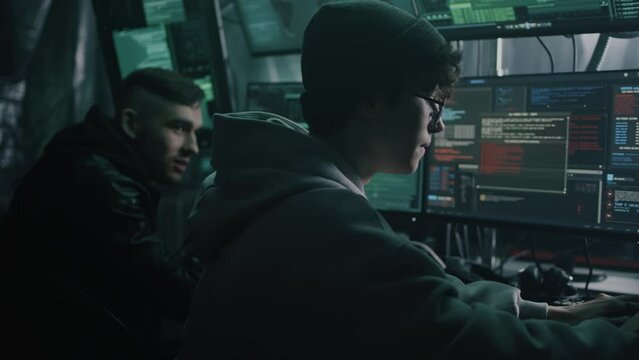 Two hackers in casual clothes cracking classified files on computer and infecting documents with a virus in a dark room with monitors