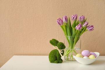 Beautiful tulips, bowl with Easter eggs and rabbit on table near color wall