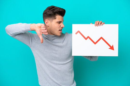 Young handsome caucasian man isolated on blue bakcground holding a sign with a decreasing statistics arrow symbol and doing bad signal