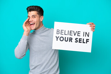 Young handsome caucasian man isolated on blue bakcground holding a placard with text Believe In Your Self and shouting