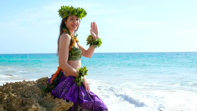 Smiling woman shakes hand saying hello.  Hula dancer posing on a tropical beach with rocks showing her hand in welcome stylized in a bikini with a flower crown in her hair.