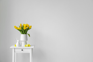 Vase with yellow tulips, Easter eggs and rabbit on table near light wall