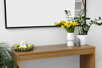 Tulips in vase, houseplant, wreath with Easter cakes and eggs on table near light wall