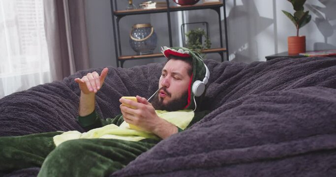 Close up portrait of adult man on sofa with smartphone in his hands listenng to music in headphones in dinosaur costume. Man chilling on couch, singing and making dacing moves. Happiness concept.